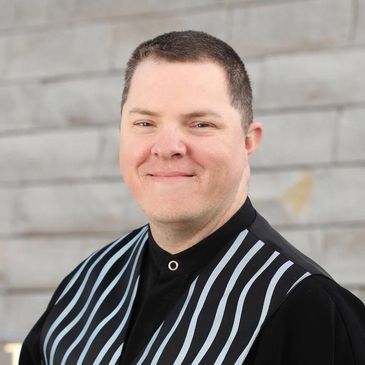 Man with short hair smiling, wearing a black shirt with vertical stripes.