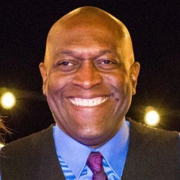 A smiling man in a blue shirt, purple tie, and black vest.