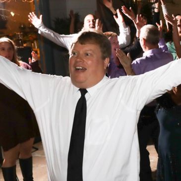 Man in white shirt and black tie joyfully leads a lively crowd with raised arms.