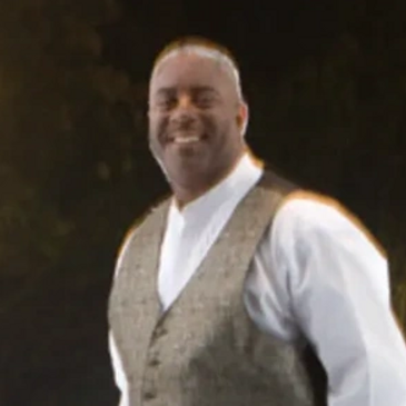 Man in a vest smiling at an outdoor nighttime event with decorative lights.