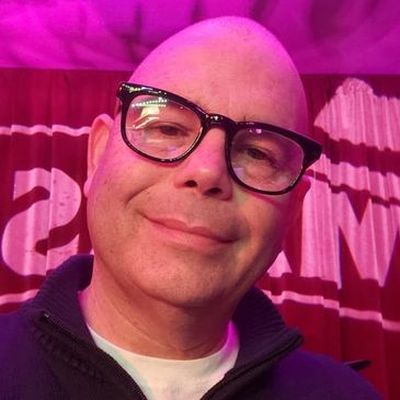 Smiling man wearing glasses and a dark zip-up sweater under pink lighting.