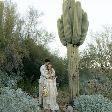 Couple in wedding attire embracing near a tall cactus in a desert setting.