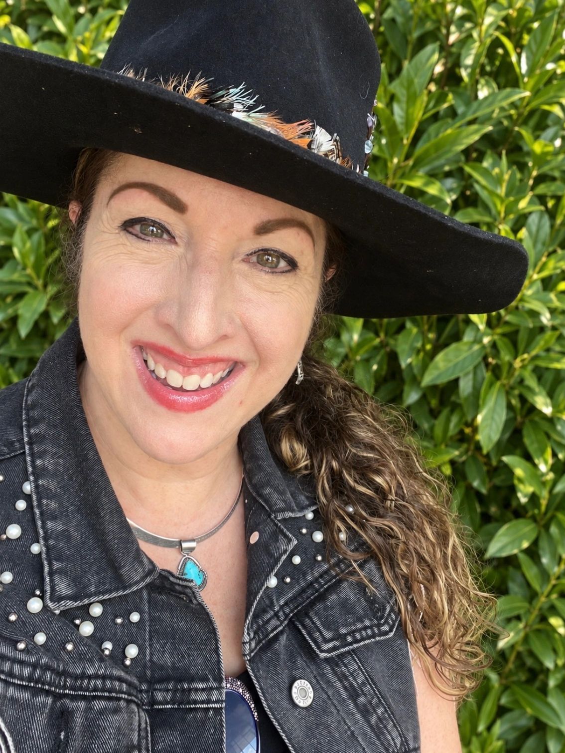 Woman smiling wearing a black hat and studded denim vest.