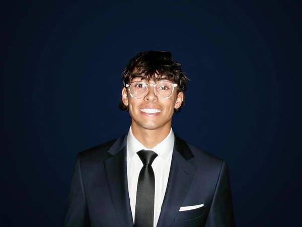 Young man in a black suit and tie smiling against a dark background.