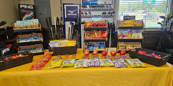 Snack display with chips, apples, and various treats on a yellow table at a golf shop.
