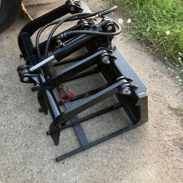 Black hydraulic grapple attachment on concrete ground near grass.