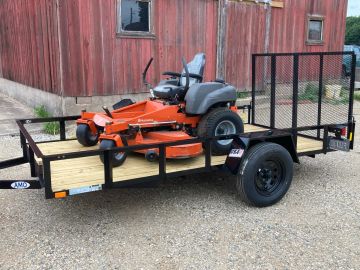 Orange Husqvarna riding lawn mower on a utility trailer outside a rustic red barn.