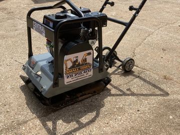 A compact plate compactor on a concrete surface with construction equipment in the background.