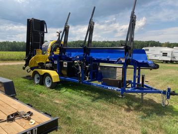 A blue wood splitter trailer with yellow accents parked on grass under a partly cloudy sky.