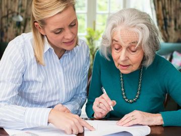 A younger woman helping an elderly woman with paperwork at a table.