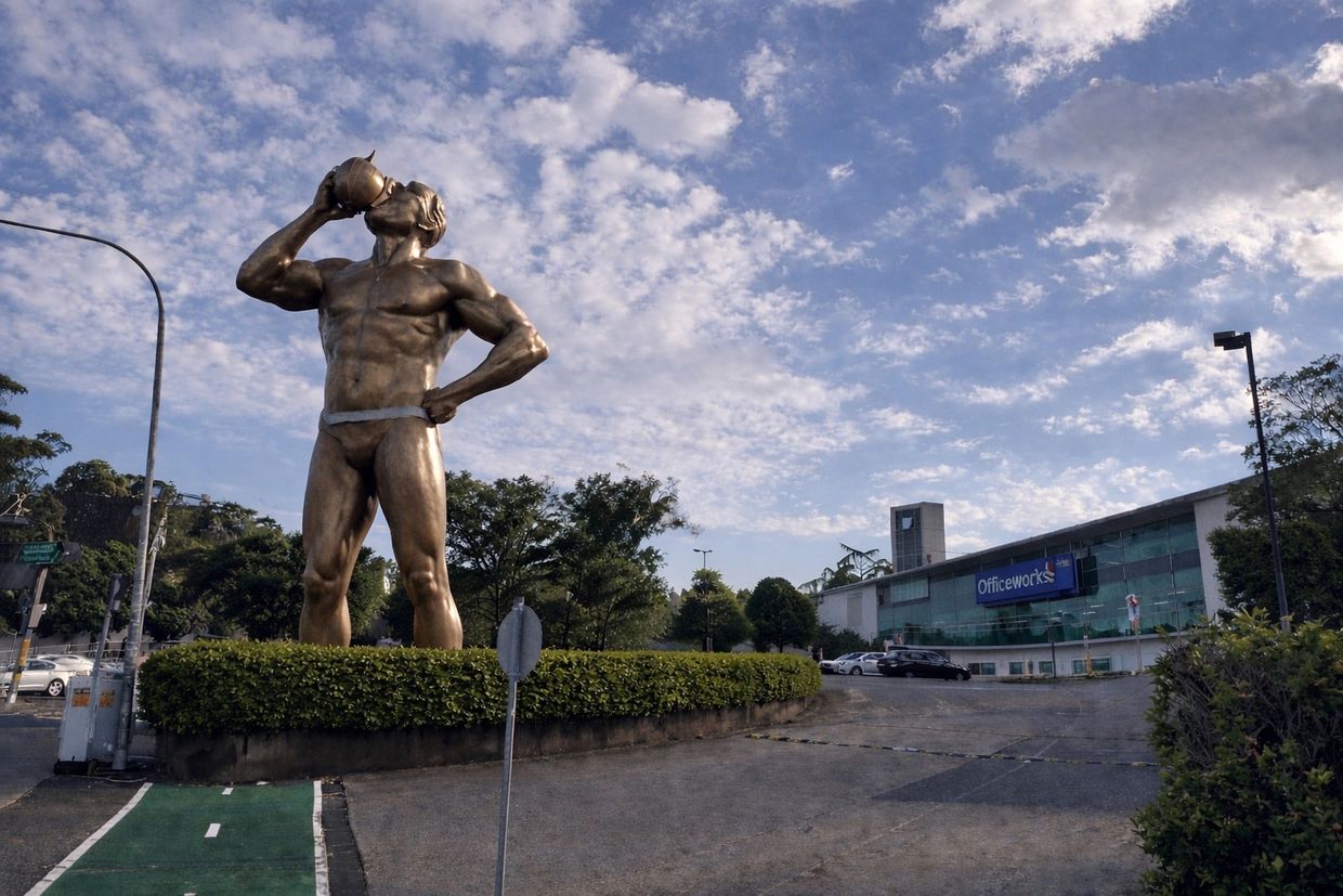 A large bronze statue of a muscular man drinking from a container outdoors.