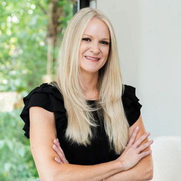 Smiling blonde woman in black top with arms crossed, standing indoors near a window.