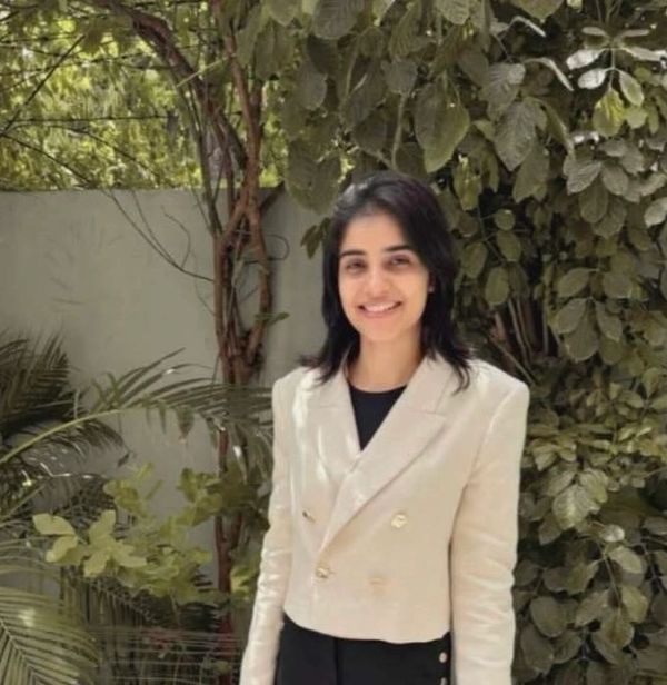 A woman in a white blazer smiling outdoors with greenery in the background.