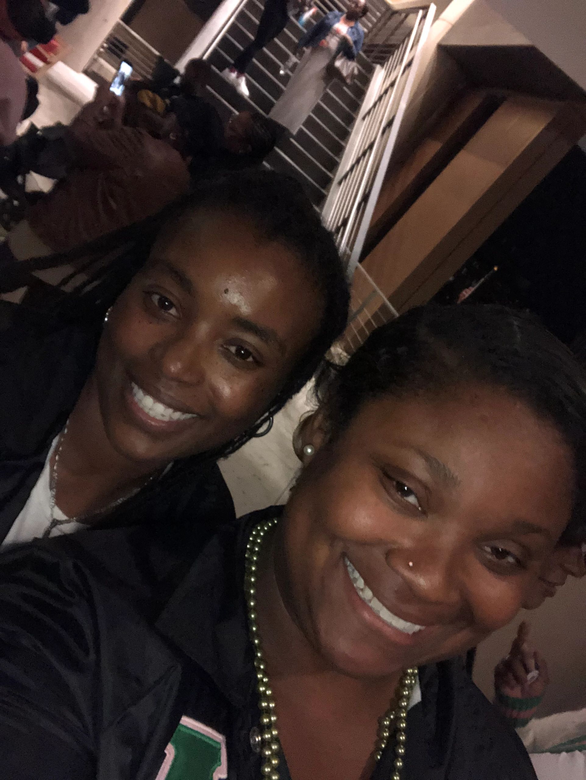 Two women smiling for a selfie in a dimly lit indoor setting with stairs in the background.