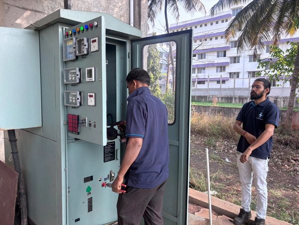 Two men working on an electrical control panel outdoors near a building.