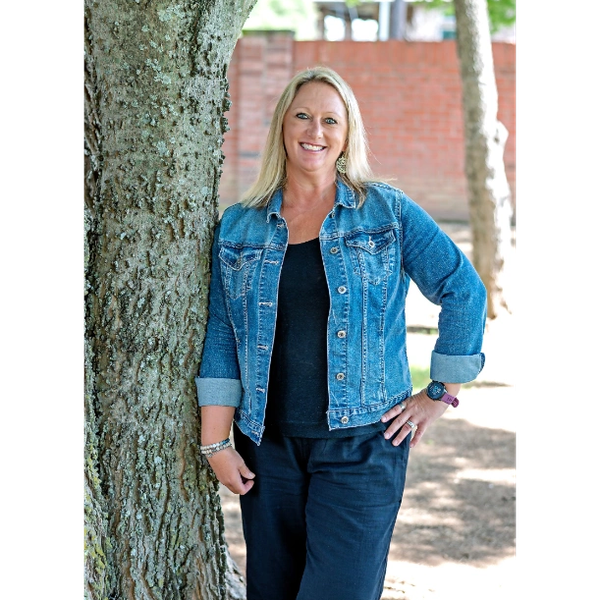 Smiling woman in denim jacket leaning against a tree outdoors.