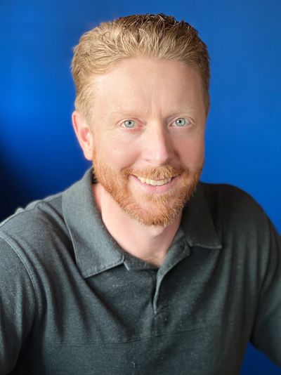 Smiling man with red hair and beard against a blue background.