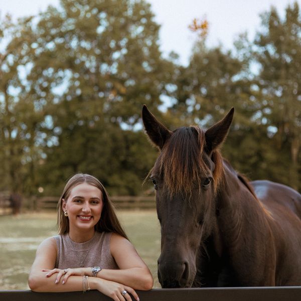 A smiling woman leans on a fence next to a dark brown horse in a serene outdoor setting.
