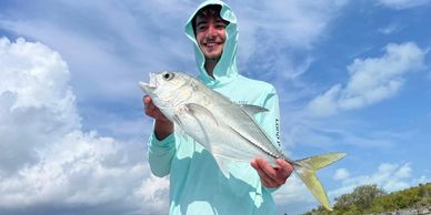 Smiling young man holding a freshly caught fish under a partly cloudy sky.