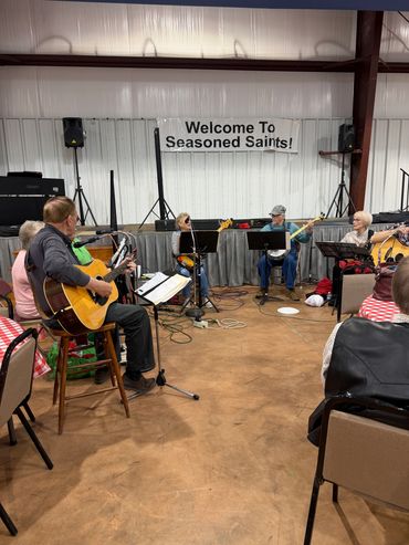A group of musicians playing guitars under a sign welcoming Seasoned Saints.