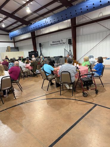 Seniors gathered in a community hall for a social event with a 'Welcome to Seasoned Saints!' sign.