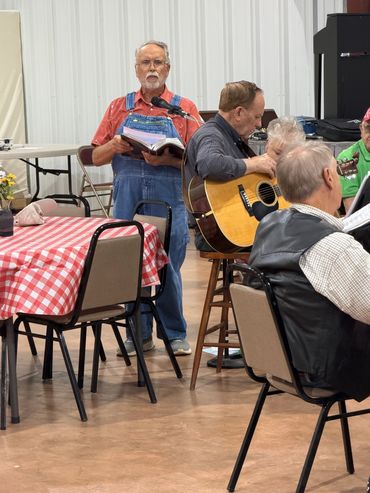 A man in overalls sings while others play guitar in a community hall.