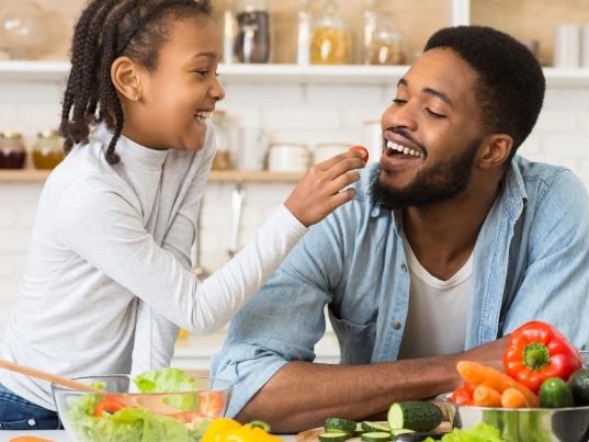 Young father being fed a piece of fruit by his 8-year old, healthy food items on the kitchen table.