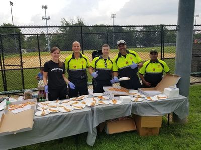 Five people, four in uniforms, serving pizza slices outdoors at a table.