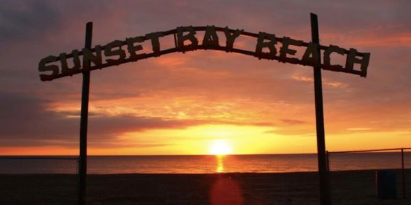 Sunset Bay Beach sign silhouetted against a vibrant sunset over the ocean.