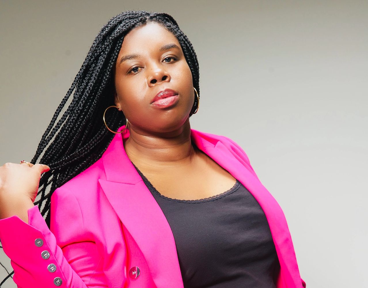 A Black woman wearing a pink blazer flicking her braids back while gazing at the camera.