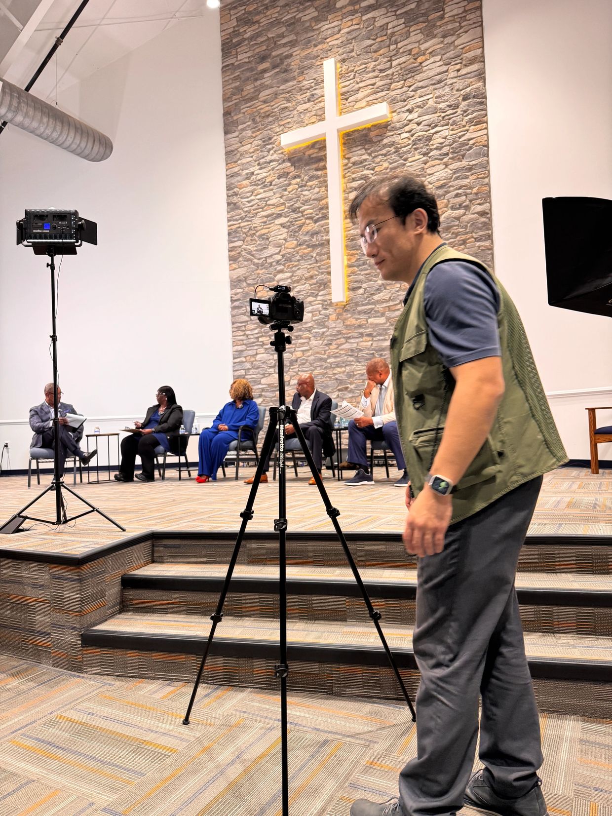 A man sets up a camera in a church with a panel discussion in progress.