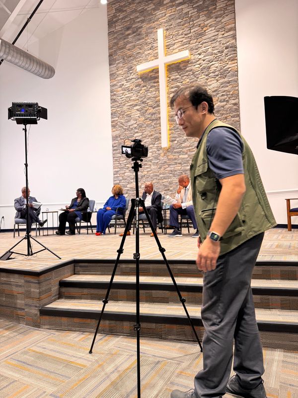 Man adjusts camera on tripod in a church setting with panelists seated on stage.