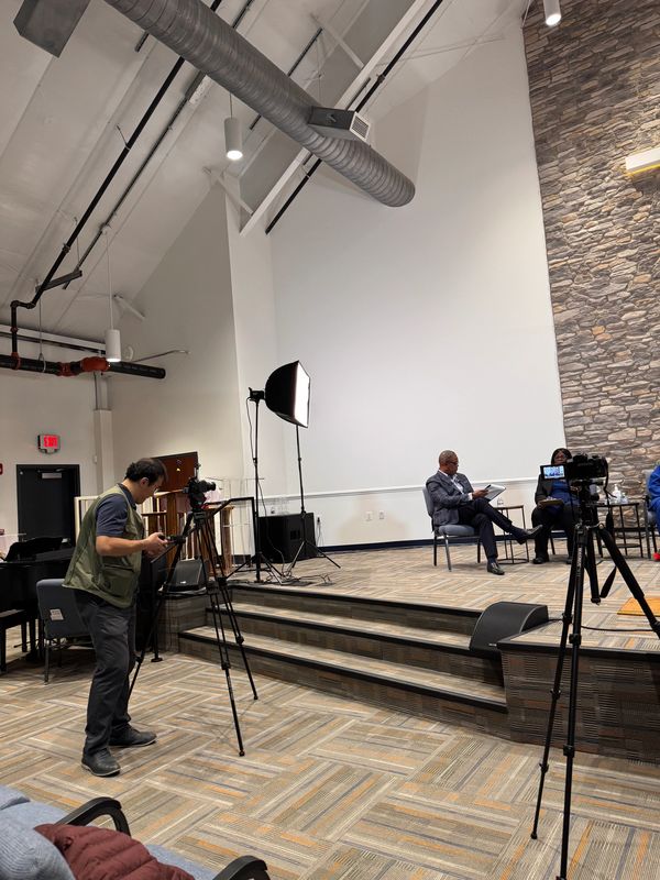A man adjusts camera equipment in a room with two seated people and a lit backdrop.