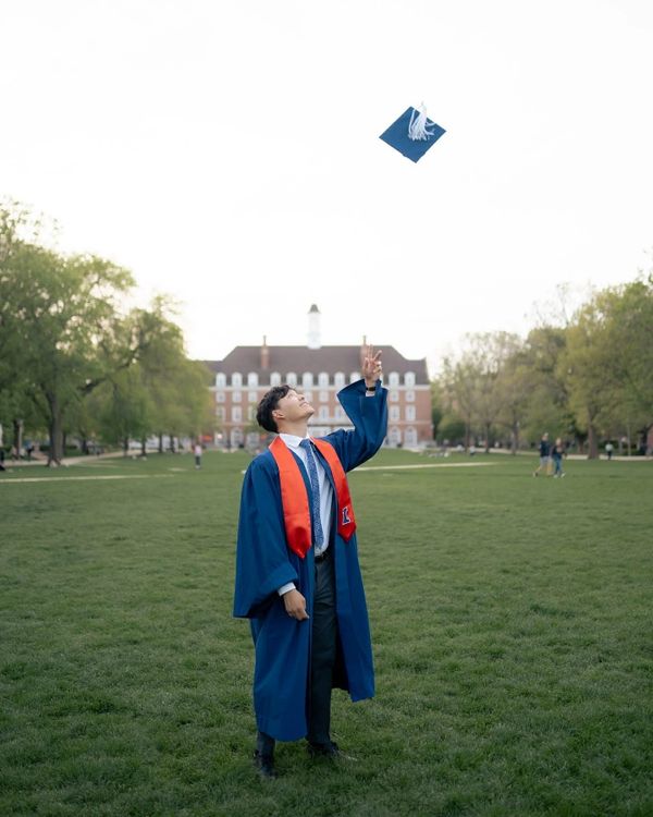 Graduate in blue gown tossing cap on campus lawn.