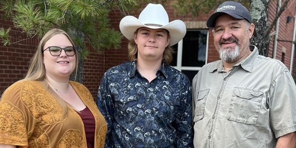 Three people smiling outdoors, including a young person in a white cowboy hat.