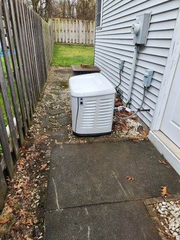 A white standby generator installed beside a house with a wooden fence and paved walkway.