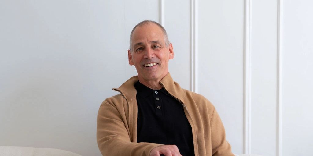 Smiling man in casual attire sitting on a white sofa against a white wall.
