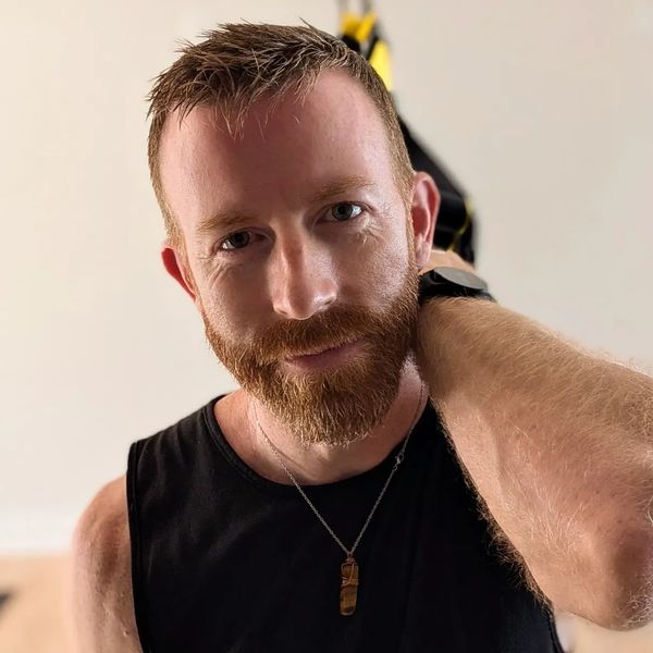 Close-up of a red-haired man with a beard wearing a black tank top and pendant.