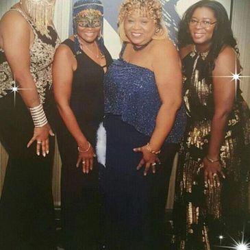 Four women dressed elegantly with decorative masks and sparkling attire at a social event.