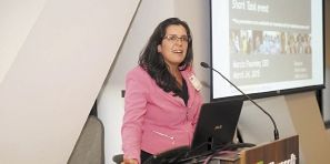 Marcia Fournier speaks at an event, with a presentation in the background.