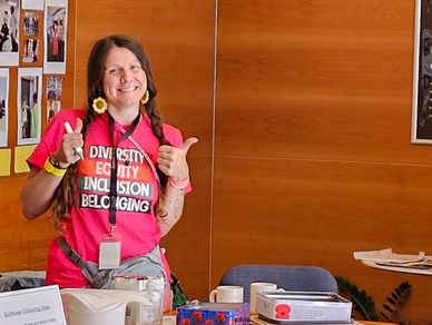 A smiling woman in a pink diversity and inclusion t-shirt gives a thumbs-up at a desk.