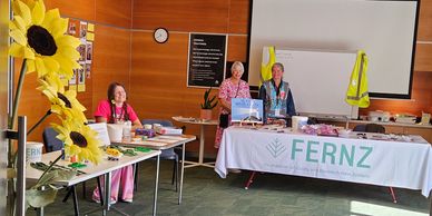 Three women at a FERNZ information booth with sunflowers and promotional materials.