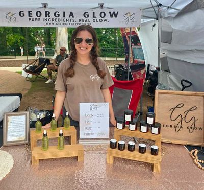 Vendor smiling at a booth selling natural skincare products outdoors.