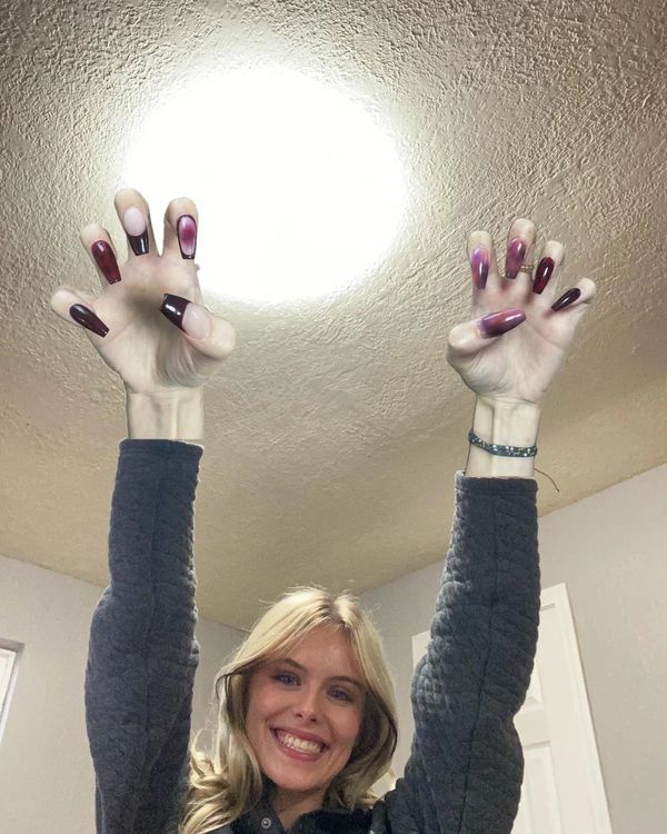 Smiling woman showing off long, burgundy-tinted nails under bright ceiling light.