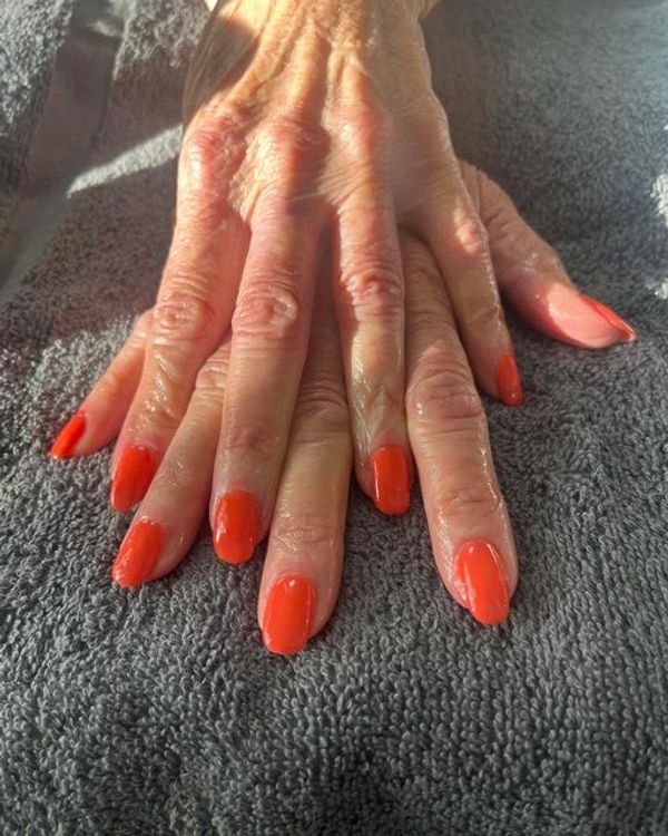 Wrinkled hands with bright orange painted nails resting on a gray towel.