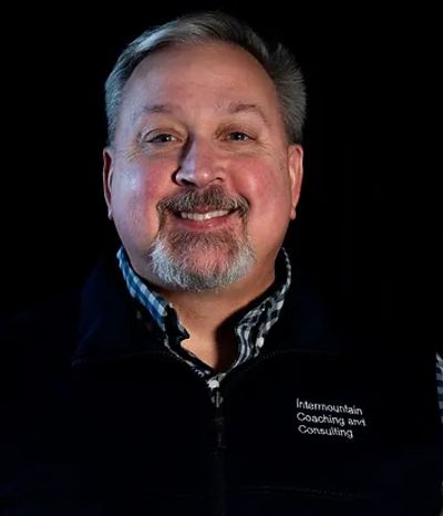 Smiling man wearing a vest with 'Intermountain Coaching and Consulting' embroidered.