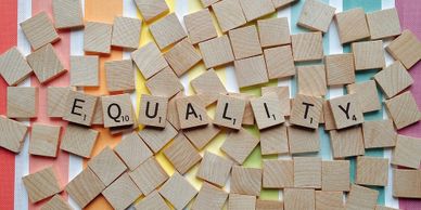 Several Scrabble tiles lay face down on a surface with rainbow and white stripes. The tiles facing upward spell out the word "Equality."