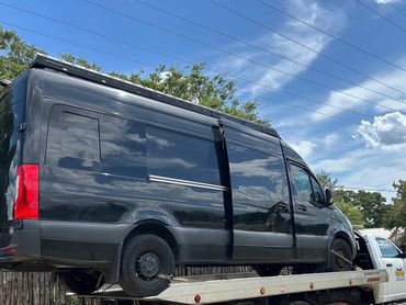 A black van is being towed on a flatbed truck on a sunny day.
