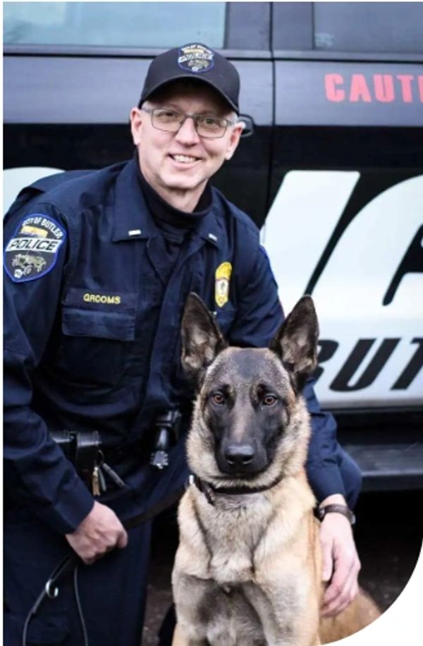 Police officer posing with a German Shepherd K9 unit in front of a police vehicle.