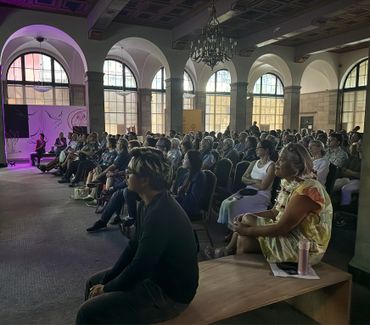 Audience attentively watching a presentation in a spacious, arched-windowed room.